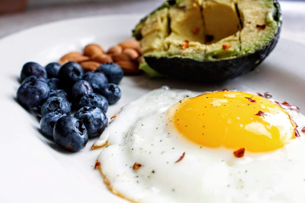 A white plate with a handful of blueberries, some almonds, an avocado, and a fried egg