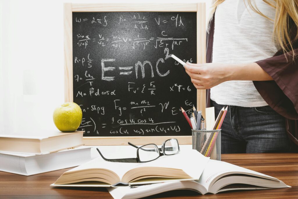 A chalkboard with E=MC squared written on it and other formulas, and books, glasses, pencils, and an apple on a desk. 