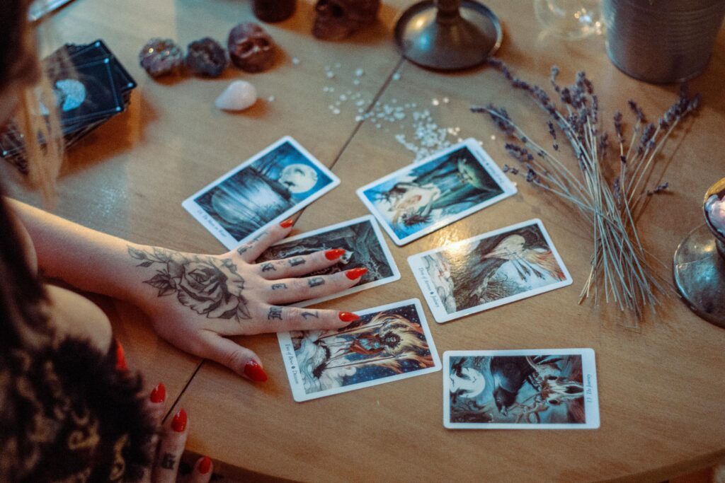 A woman with tattooed hands reads tarot cards on a table. The cards are surrounded by crystals, candles, and lavender