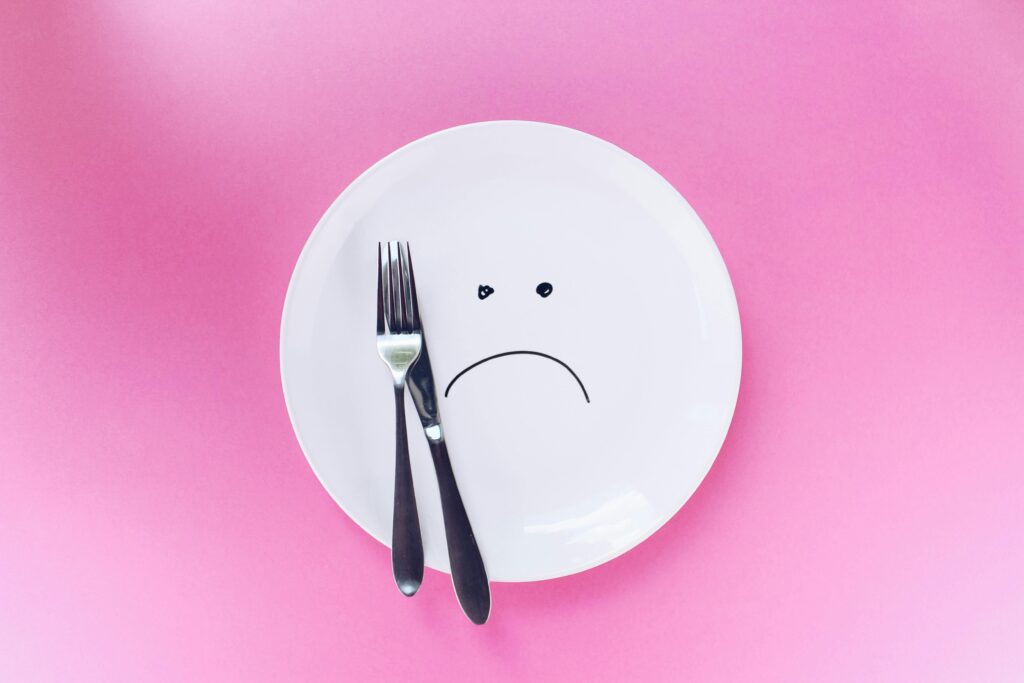 A white plate on a pink background. There is a knife and a fork on the plate. The plate has a sad face drawn on it.