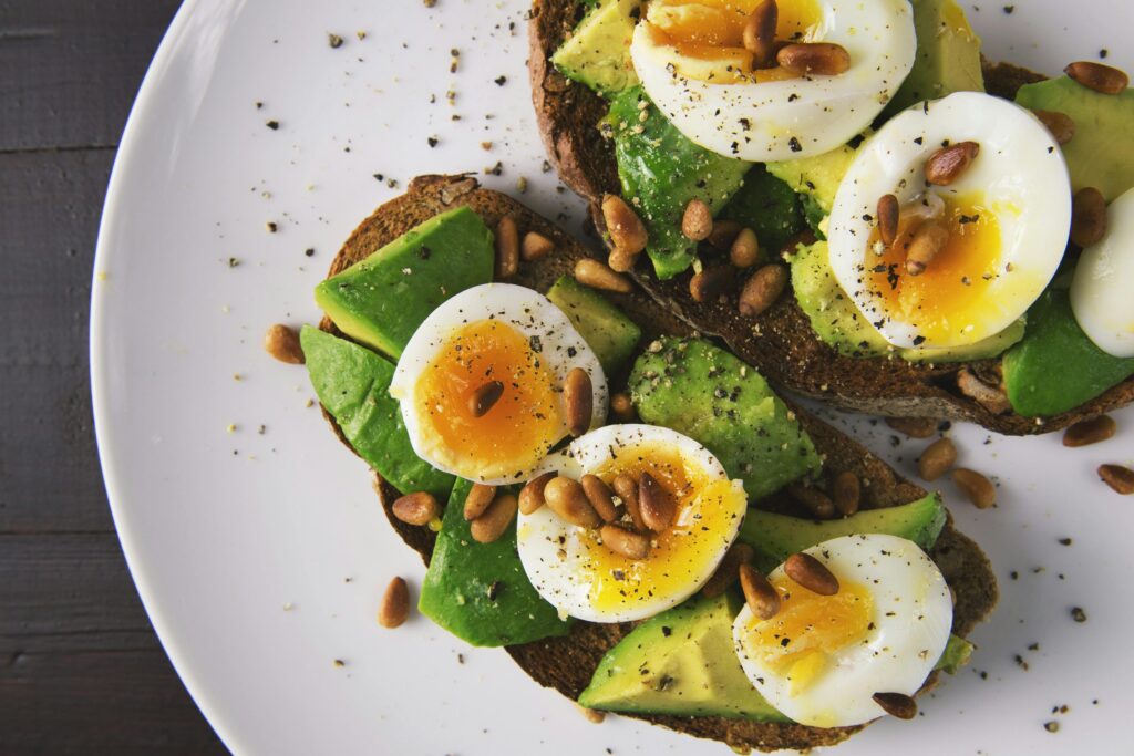 Toast on a white plate, topped with avocado, boiled eggs, and pine nuts