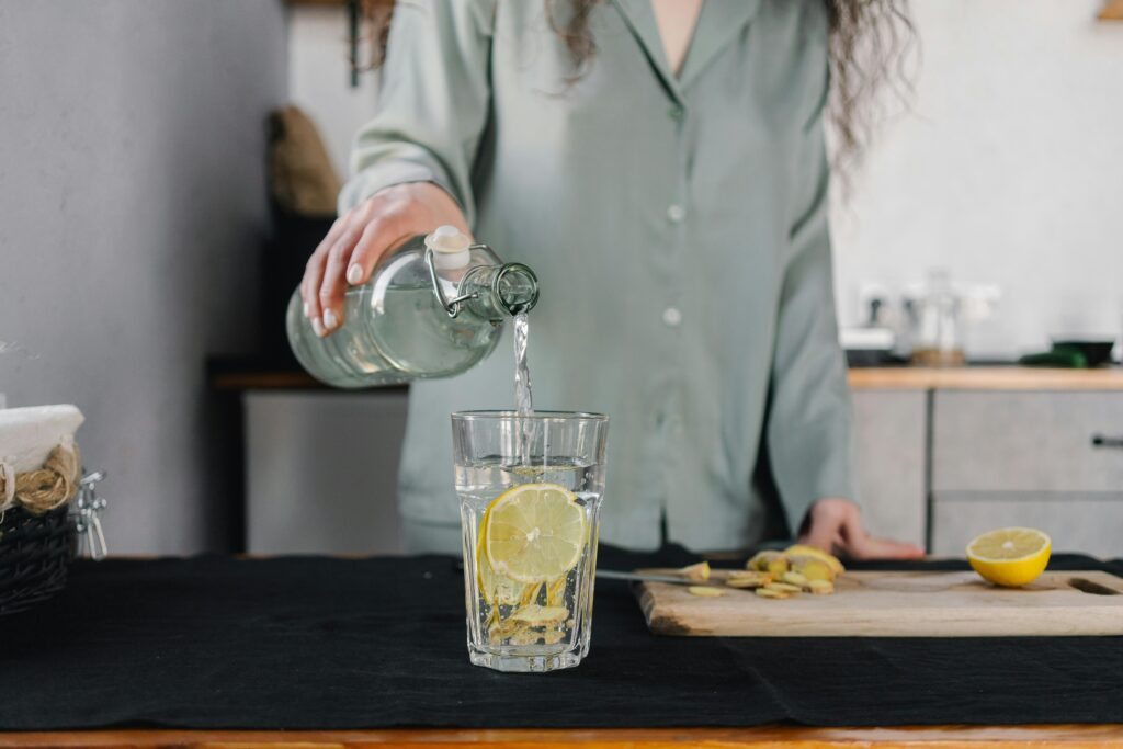 A woman in a grey shirt pouring water from a bottle into a glass filled with slices of lemon
